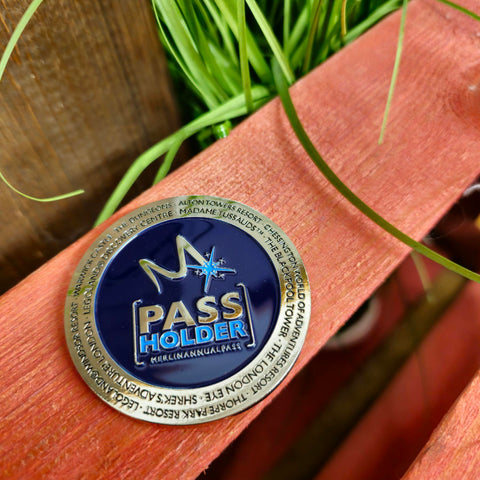 A photograph of a round metal magnet, it has the "m" symbol in the middle with the words "pass holder" underneath it on a royal blue background. Around the outside is a silver metal circle with the names of the Merlin Attractions on it in the same dark blue colured text
