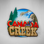 A shaped wooden sign with the Canada Creek logo on the top surface. The logo has the word Canada in red text, Creek in a light tan stone colour and the background is a natural scene with two pine trees and a waterfall.