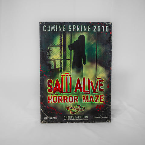 A portrait orientated poster with 'Coming Spring 2010' at the top in a very light green font. SAW Alive Horror Maze logo in the lower half and an image of a shadowy figure behind some fencing and industrial equipment as the main image.