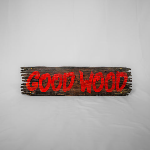 A wooden sign with the words Good Wood lettered on it in red paint. The ends are jagged as if it hasn't been hewn properly.