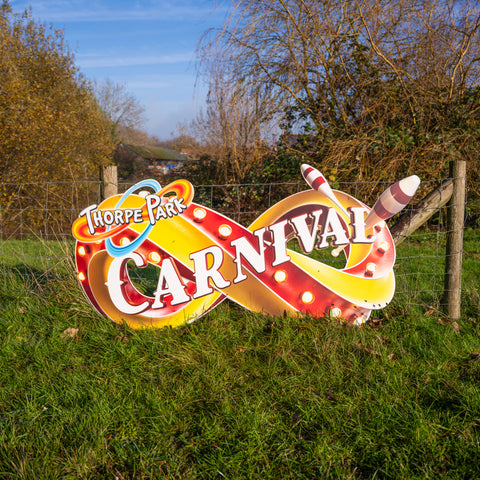 A shaped sign in landscape orientation. It is of an infinity look in red and yellow to look like a circus sign. There are two juggling batons on the right hand side. To the left is the old Thorpe Park logo and along the middle in white text with a red outline is the word Carnival