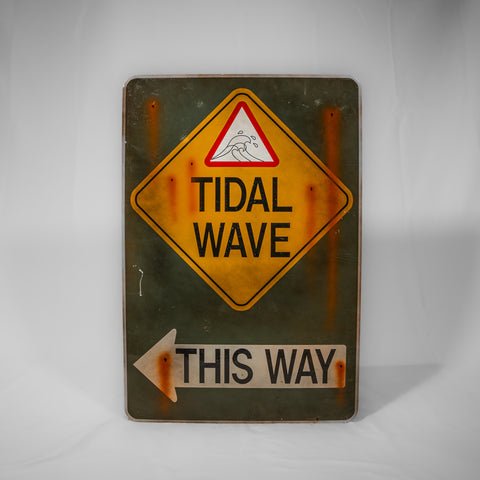 A portrait orientated metal sign, it is an army green colour with a yellow hazard sign with the words Tidal Wave in at the top. Underneath is a white arrow pointing to the left with This Way written in it in black text. The whole sign has printed on rust marks as if there is water damage