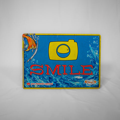 A landscape orientated sign which is blue in the middle with water effect across the bottom. There is a stylised image of a camera in yellow at the upper middle, and the word "smile" in red text underneath it. In the top left corner is an orange fish tail.