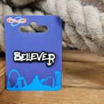 A black pin badge with a slightly off-white infill which says the word Believer in a graffiti style.