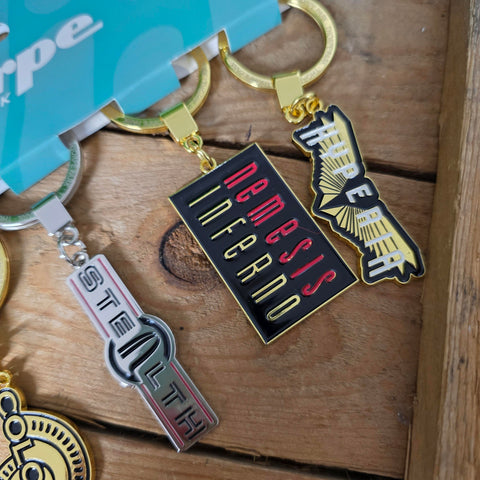 Metal keychains with 'Nemesis Inferno' and 'Stealth' designs on a wooden surface.