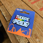 A photograph of a pin badge, it is cut to the shape of the Thorpe Pride logo and is a dark metal base with coloured inlay