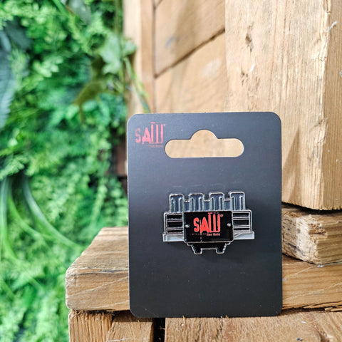 A pin badge in the shape of the front of the SAW - The Ride ride vehicle. It is rectangular in black, with four seat tops along the top in dark grey. In the middle is the SAW - The Ride logo in red.