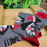 Grey socks, with the Billy doll face and red spirals across them. The SAW - The Ride logo is on the sole in black text