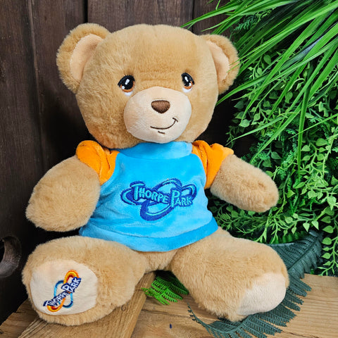 A light brown teddy bear, wearing a light blue and orange sweatshirt. The old Thorpe Park logo is embroidered on the sweatshirt and his foot