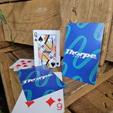 A photograph of playing cards, the backs of which are blue with a teal coaster track pattern and the Thorpe Park logo in white in the centre. The fronts are a standard deck of cards.