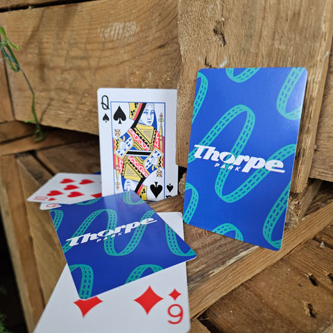 A photograph of playing cards, the backs of which are blue with a teal coaster track pattern and the Thorpe Park logo in white in the centre. The fronts are a standard deck of cards.