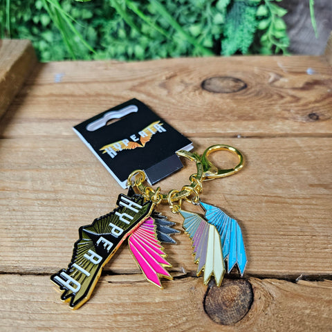 A keyring with multiple charms, one is the Hyperia logo of gold wings and white 'Hyperia' text. There are then four other single wings, which are in pink, grey, blue and cream with green. The keyring loop is gold and there is a clip also in gold next to the charms.