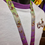 A lanyard on top of a white t-shirt, it is purple fading up to green and features the mardi gras logo in different sizes and angles across it.