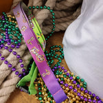 A photograph of a lanyard, it is purple fading into green, and thas the Mardi Gras event logo scattered across it at different sizes and angles. Behind it are some beads and rope.
