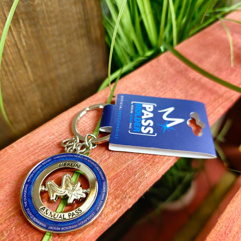 A photograph of a round keyring, it has the letter "m" in the middle with the names of all attractions around the outside on a dark blue circle, inside that is a silver metal circle with the words "Merlin Annual Pass" embossed into it.
