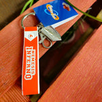 A photo of the keyring, it has an orange strap with the words Nemesis Inferno on it, and two charms - a metal clip embossed with the logo and a keyring clip
