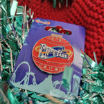 A photograph of a pin badge. The shape is circular and red with two glittery red bands at the top and bottom.  There's a Thorpe Park logo in the middle with some glittery frost icicles added in white. Below the pin badge is light blue tinsel and in the background a red knitted santa hat.