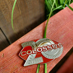 A photograph of a maroon coloured oval shaped magnet with a large silver 20 in the background. The world Colossus is across the middle in silver.