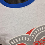 A photograph of a white t-shirt with royal blue collar and sleeve hems, in the middle is the Colossus rollercoaster logo with a silver 20 behind it.