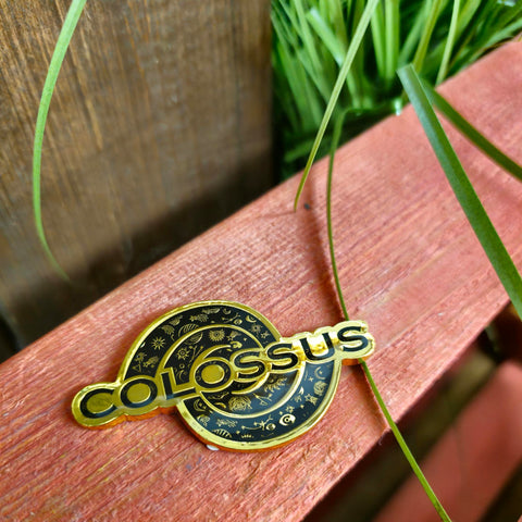 A photo of the Colossus logo which is the word colossus with a spiral behind it. The borders and text outline is in gold, and the letters and background of the spiral is a dark navy blue. Amongst the spiral are images of stars and astrology symbols
