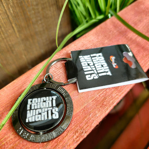 A photograph of the keyring, it is round with an outer circle in a grey metal. The inner circle is black with the Fright Nights logo on it.