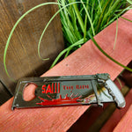 A photo of the magnet, it is a silver hacksaw shape with a bottle opener on the left hand side, in the middle is the SAW - The Ride text in red, and underneath is a blood splatter, also in red.