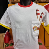 A photograph of the t-shirt, it is mostly white, but the collar detail is bright red, and the sleeve detail is yellow. There is a pocket on the chest area with the carnival logo printed on it, and some juggling batons printed to look as if they are coming out of the top.