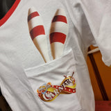 A close up of the pocket detail of the t-shirt, there is a carnival logo printed on top of the pocket, and there are two juggling batons printed as if they are coming out of the top. You can also see the red collar at the top of the photo. The main t-shirt is white.