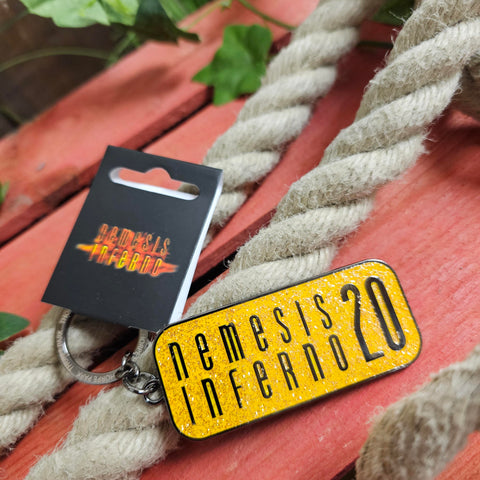 A keyring shaped like a rectangle with rounded corners. The middle is yellow with orange glitter and the words Nemesis Inferno 20 are in black in the middle