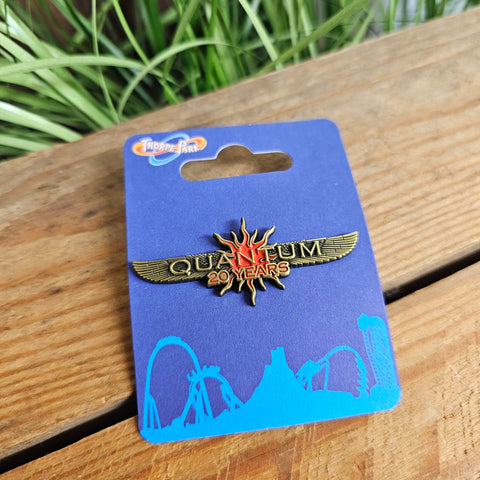 A pin badge shaped like two outstretched wings, it is an off-gold colour with a red sunburst in the middle. The word Quantum is raised in the middle in gold, with 20 Years underneath in red.