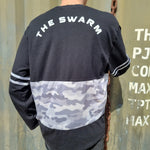 A photograph of the back someone wearing a t-shirt, it is black at the top, then halfway down switches to a camouflage print in grey tones, then a final band of black around the base. Across the shoulders is the wording "The Swarm" in white text. The sleeves have two thin (around 1cm) camo print bands stitched on which match the inserted panel on the main body. The person is in front of a green metal wall with a chain and some text to the right hand side.