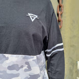 A close up photograph of the embroidered symbol on the t-shirt. It is a light grey colour and is a triangle with a horizontal line through it. You can also see where the camouflage panel is stitched in, and the striping on the arms.