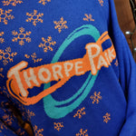 A photograph of a knitted jumper, it is in a royal blue with orange detailing on the collar and cuffs. There are orange snowflakes embroidered into the right hand shoulder. In the middle is a Thorpe Park logo.