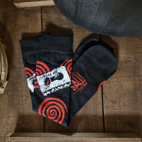 A photograph of a pair of socks, they are black with red spirals randomly across them, and a white cassette tape which has "play me" written on it around the ankle area. On the sole is the SAW - The Ride logo. The socks are on a dark wooden surface.