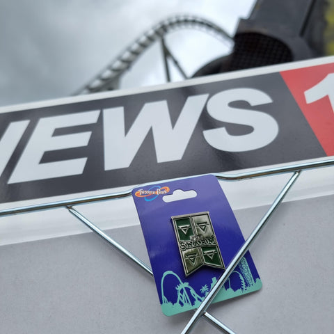 A photograph of the pin badge trapped to the News 16 billboard. Behind it is the rollercoaster "The Swarm"