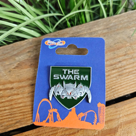 A pin badge shaped like a shield, it is army green with the words The Swarm in silver at the top and a silver outline of the Swarm ride vehicle