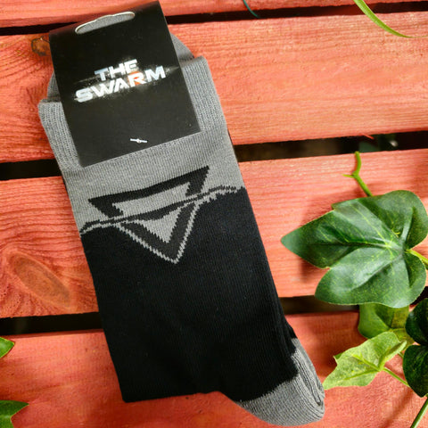 A pair of socks folded in their packaging, the socks are black with grey details on the ankles and heel. The Swarm triangular logo is on the ankle area