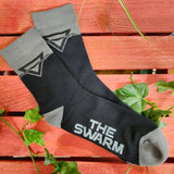 A pair of socks unfolded, the socks are black with grey details on the ankles and heel. The Swarm triangular logo is on the ankle area and the words The Swarm on the sole