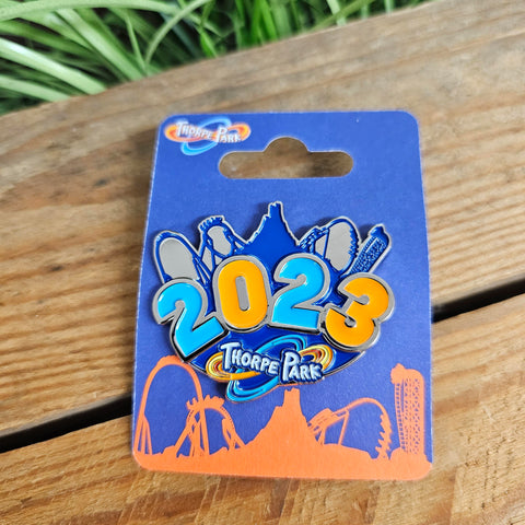 A photo of the 2023 pin badge, it has the skyline in navy blue across the top with an arched 2023 text underneath in alternating blue and orange lettering. Underneath is a Thorpe Park logo