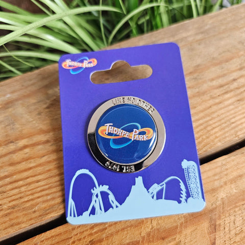A photo of the pin badge, it is round and silver, with a blue Thorpe Park logo in the middle. Embossed around the outside is "like no other" and "est 1979"