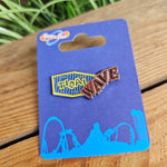 A pin badge shaped like the entrance sign for Tidal Wave. The word 'Tidal' is in yellow on a blue squashed diamond shape, and the word wave is in red to the right looking like neon light lettering