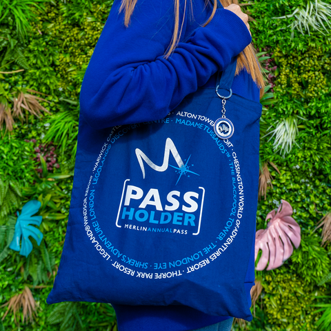 A photograph of someone holding the shopper bag, it is royal blue and has the words "pass holder" in the middle, just below a silver letter "m". In a circle around this is a list of the Merlin Attractions in light blue, with an outer circle containing more attraction names in white.